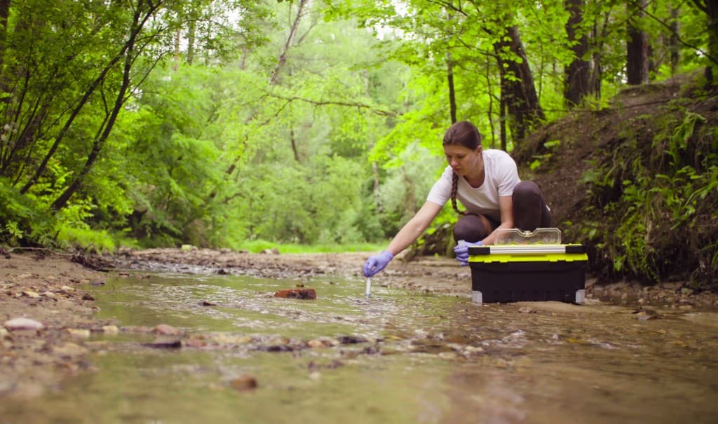 Technician wearing gloves collects a water sample from a shallow stream in a forest beside field monitoring equipment.